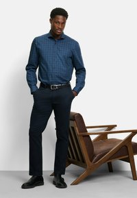 Marine-kariertes Hemd mit langen Ärmeln, dunkle Hose und schwarze Schuhe, steht neben einem braunen Holzstuhl.