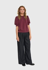 Mujer de pie con cabello rizado, vistiendo una blusa de mangas abullonadas burdeos, pantalones anchos negros y zapatos negros, frente a un fondo liso.