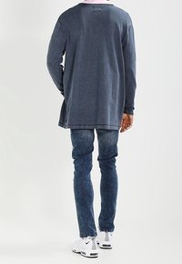 Dunkelblauer Oversized-Sweatshirt mit langen Ärmeln, rundem Halsausschnitt und rohen Kanten, kombiniert mit dunkelblauer Jeans und weißen Turnschuhen.