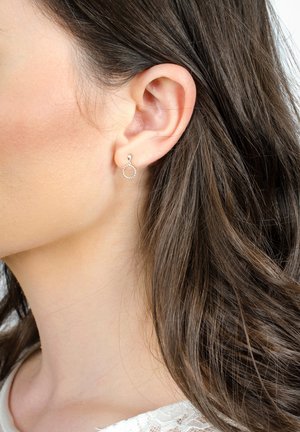 Silverfärgade hoop-örhängen med en texturerad, oregelbunden kant, som visar en cirkulär design. Bärs på en models öra, tillsammans med en spetsblus.