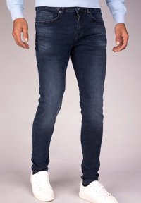 Hombre vestido con jeans oscuros ajustados y zapatillas blancas, de pie frente a un fondo neutro.
