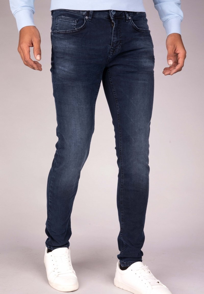 Hombre vestido con jeans oscuros ajustados y zapatillas blancas, de pie frente a un fondo neutro.