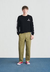 Νέος άνδρας στέκεται με τα χέρια στις τσέπες, φορώντας μαύρο φούτερ Converse, χακί παντελόνι και κόκκινα αθλητικά παπούτσια σε μπλε πάτωμα.