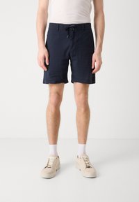Bermudas azul marinho com cintura elástica e bolsos laterais, feitas de um tecido texturizado, combinadas com ténis de camurça bege e meias brancas.