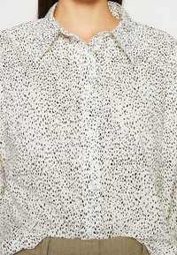Chemise blanche boutonnée avec des points noirs irréguliers portée rentrée dans un pantalon marron clair, montrant le col et le haut du torse.