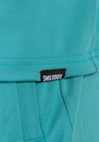 Türkisfarbener Stoff mit glatter Textur. Verfügt über ein schwarzes Etikett mit der Aufschrift "SMILODDY". Sichtbare genähte Nähte und ein einfaches, modernes Design.