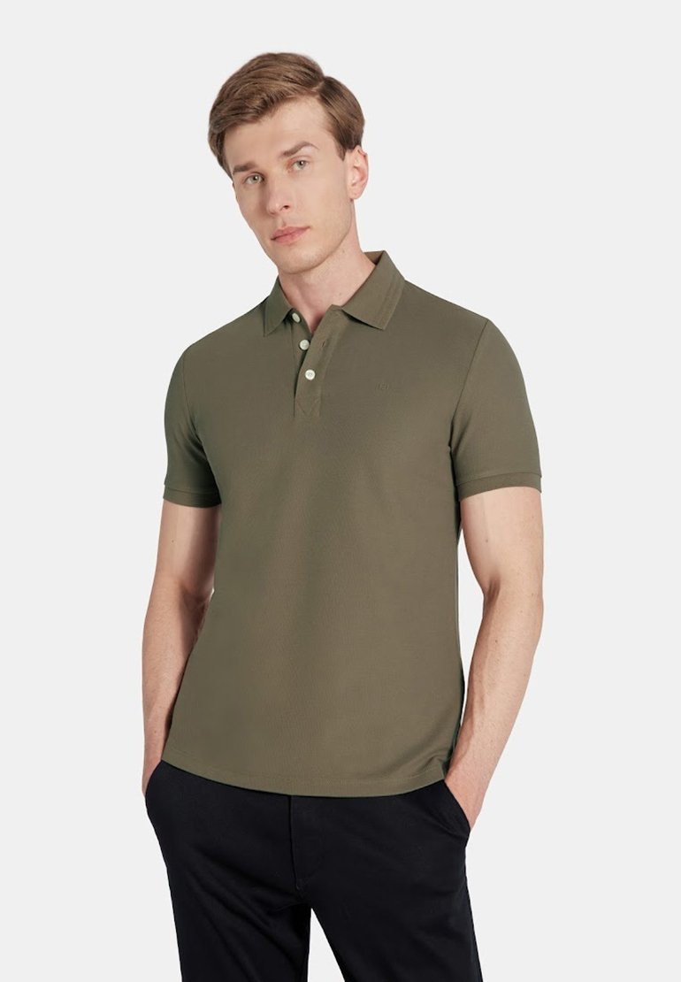 Oliwkowa koszulka polo z teksturowanej tkaniny, z klasycznym kołnierzykiem i zapięciem na trzy guziki; zestawiona z ciemnymi spodniami.