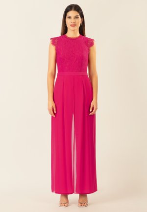 Frau mit langen dunklen Haaren, die ein ärmelloses Spitzenoberteil in Fuchsia und weit geschnittene Chiffonhosen trägt, steht vor einem schlichten hellen Hintergrund.