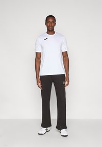 Man standing facing forward wearing a white sports t-shirt, black sweatpants, and white athletic shoes against a plain background.