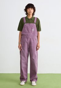 Gli salopette di jeans viola presentano spalline regolabili, bottoni laterali e tasche frontali, indossate sopra una maglietta polo in maglia verde. Il materiale sembra morbido.