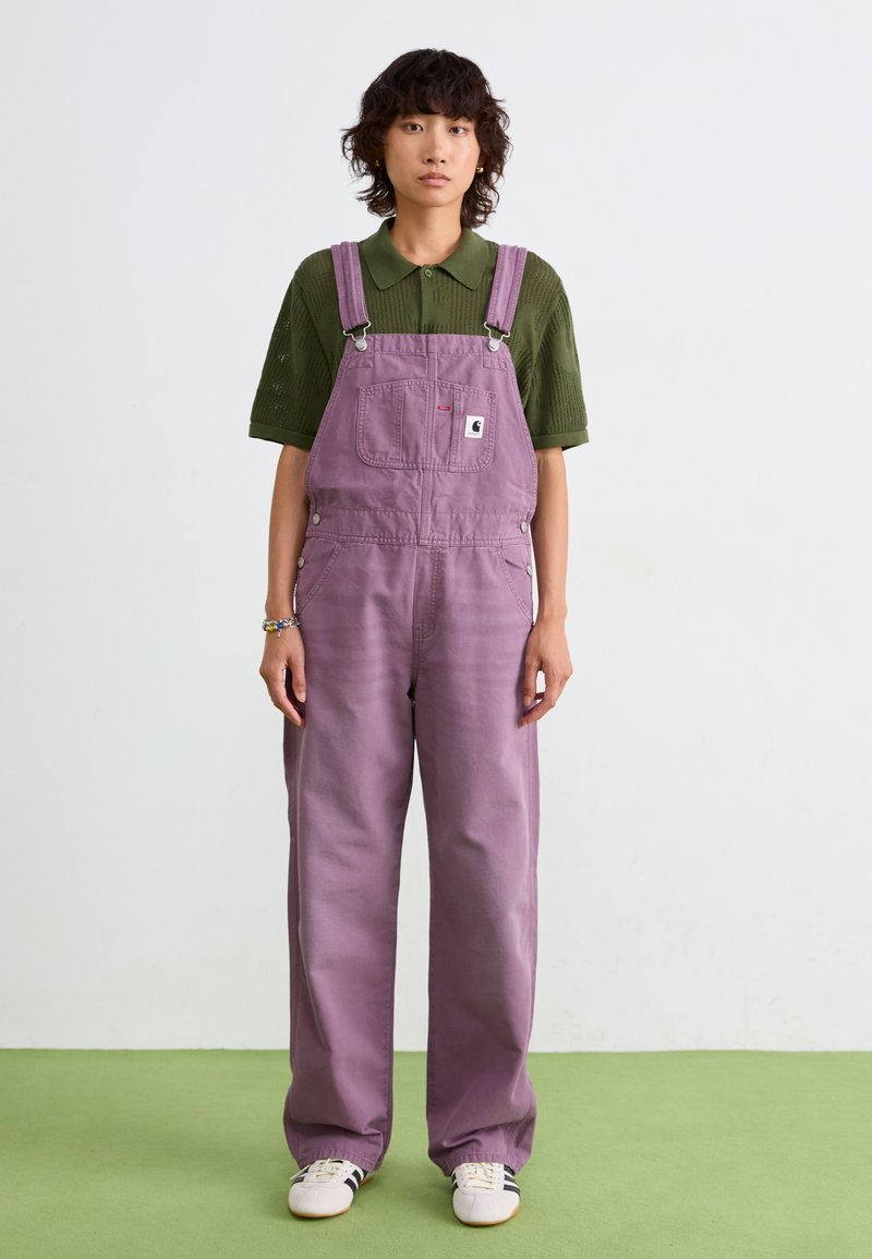 Gli salopette di jeans viola presentano spalline regolabili, bottoni laterali e tasche frontali, indossate sopra una maglietta polo in maglia verde. Il materiale sembra morbido.