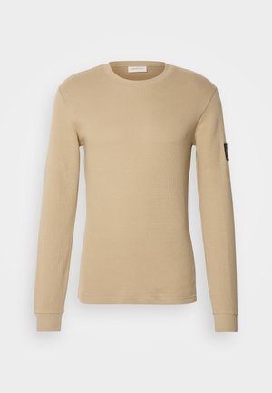 Long-sleeve, beige T-shirt made of textured fabric, featuring a round neck and fitted cuffs. Includes a small black logo on the sleeve.