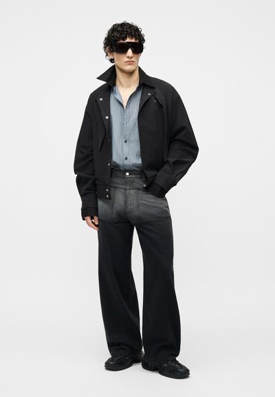 Mann mit schwarzer Sonnenbrille, schwarzer Jacke über einem blau gestreiften Hemd, dunkler weiter Hose und schwarzen Sneakern, stehend vor weißem Hintergrund.