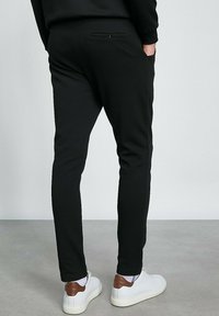 Pantalons de jogging noirs avec une coupe slim, taille élastique et poche zippée au dos, associés à des baskets blanches avec des accents marron. Texture lisse.
