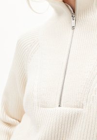 Pull-over blanc en maille avec un col montant, doté d'une fermeture éclair argentée et d'un bord-côtes texturé. Comprend un détail de poche avant.