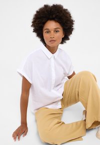 Camisa blanca de manga corta con cuello abotonado y un logo sutil, combinada con pantalones sueltos de color beige claro, todo sobre un fondo liso.