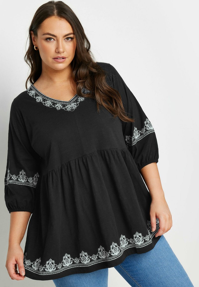 Tunic nera con scollo a V e motivi floreali ricamati in argento. Presenta maniche a sbuffo e orlo arricciato, abbinata a jeans blu.