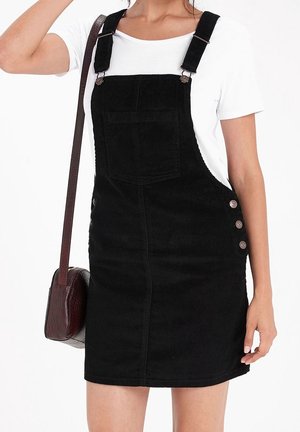 Donna che indossa un vestito nero di ciniglia a salopette sopra una maglietta bianca, con una borsa a tracolla marrone testurizzata, su uno sfondo bianco.