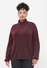 Suéter de punto burdeos con cuello alto, que cuenta con un cierre de cremallera y un ajuste ligeramente holgado. Textura suave y patrón acanalado en toda su superficie.