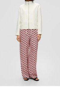 Giacca con collo arrotondato bianco e chiusura con zip. Pantaloni larghi rosa con un motivo a cuori ripetuto in rosa scuro. Sandali neri.