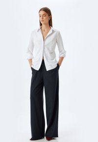 Camicia bianca con maniche arrotolate, abbinata a pantaloni a gamba larga a righe sottili scure. Tessuto morbido con dettagli minimi.