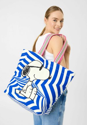 Frau trägt weißes Tanktop und Jeans und eine große blau-weiß gestreifte Tragetasche mit Snoopy-Grafik und pink gestreiften Griffen.