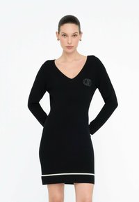 Donna che indossa un vestito lungo nero aderente con scollo a V, piccolo logo ricamato sul petto e striscia bianca vicino all'orlo, in piedi con le mani dietro la schiena.