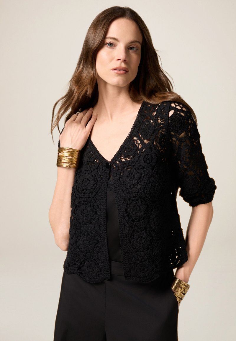 Svart virkade cardigan med blommönster, korta ärmar och knäppning, bärs över en svart topp. Har ett guldarmband.