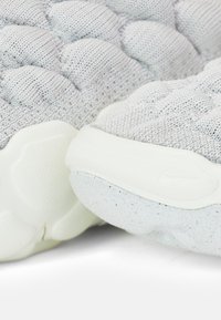 Nära bild av ljusgrå stickade sportskor med texturerade vita sulor och prickigt greppmönster.