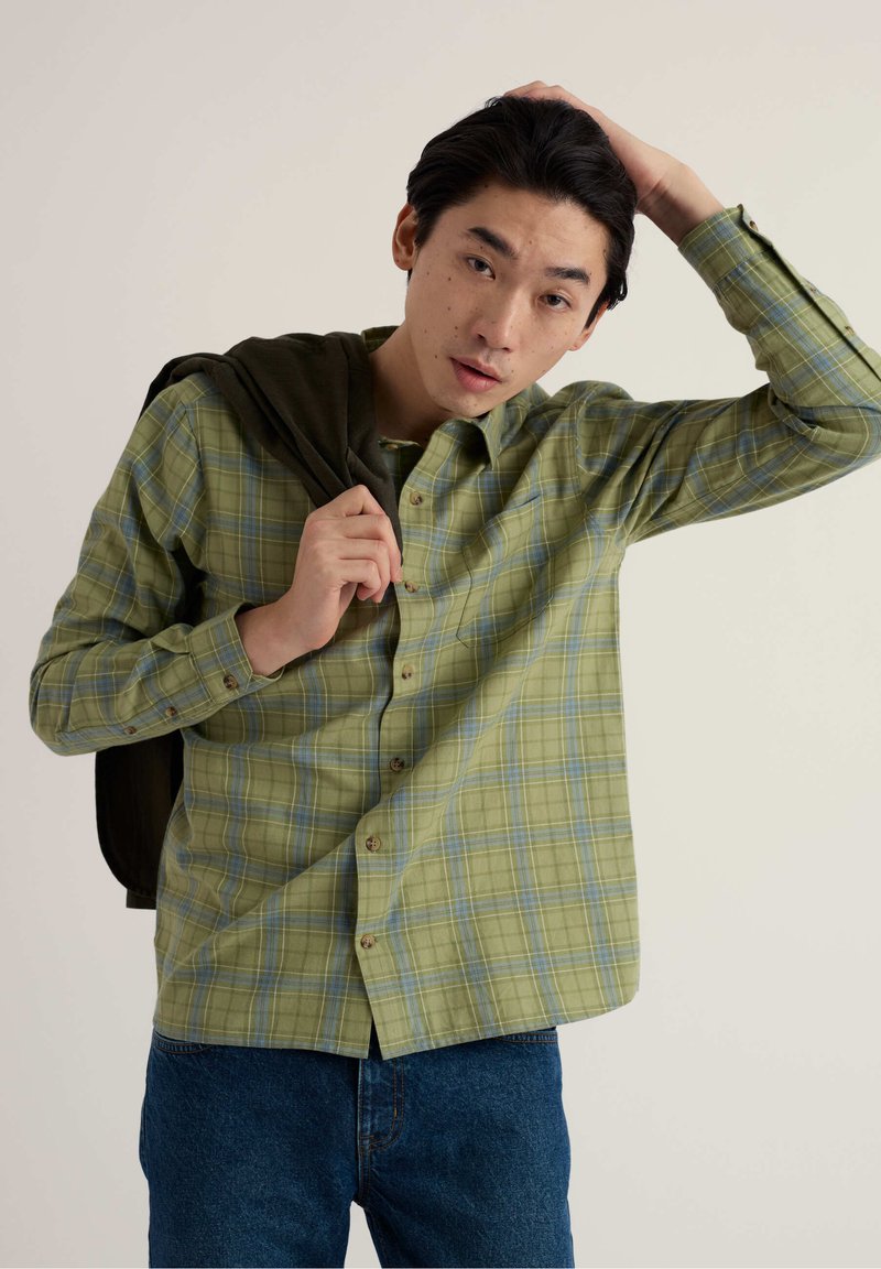 Jeune homme portant une chemise verte à carreaux et un jean bleu, tenant une veste sombre sur une épaule et touchant ses cheveux de l'autre main.