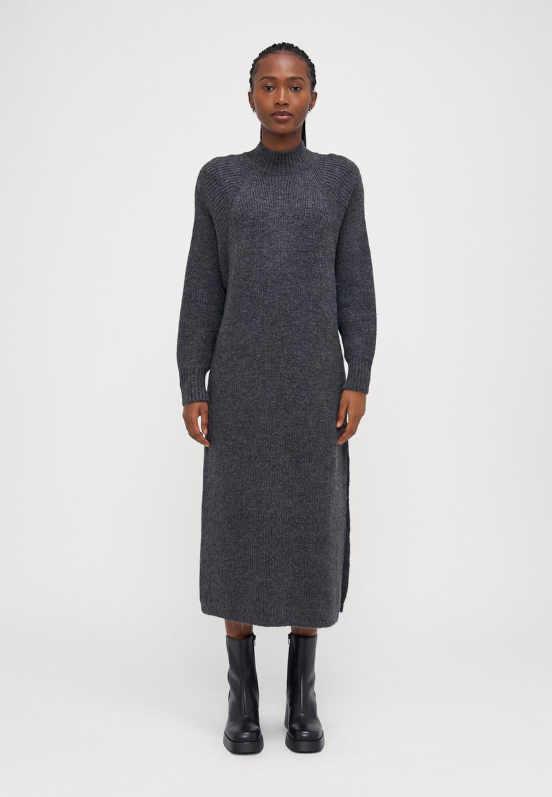 Μακρύ, σκούρο γκρι πλεκτό φόρεμα με ψηλό λαιμό και ριμπ λεπτομέρειες. Διαθέτει πλαϊνές σχισμές και συνδυάζεται με μαύρες μπότες μέχρι το γόνατο.