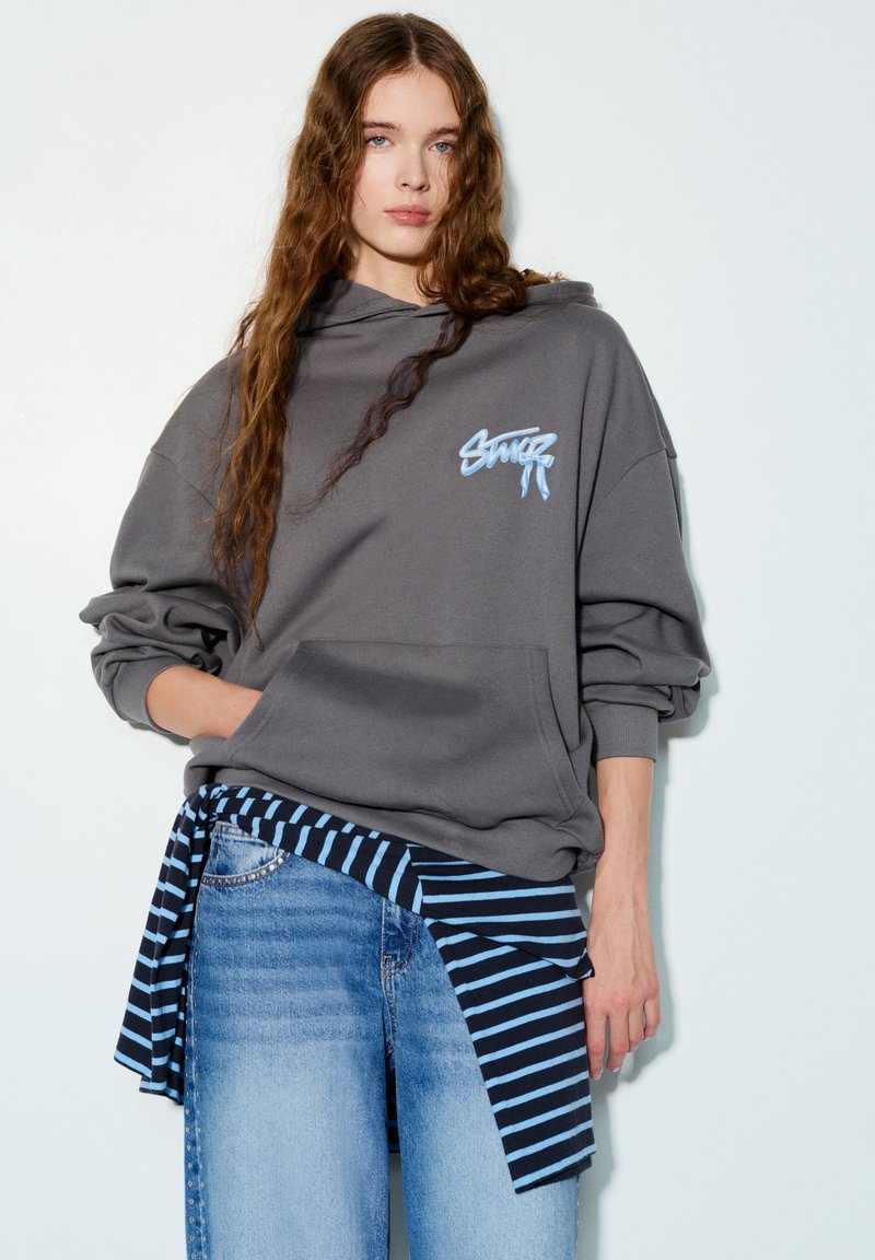 Siva oversized majica s kapuljačom i džepom sprijeda s plavim logotipom. Uparena s plavim trapericama i vezanom tamnoplavo-prugastom košuljom oko struka.