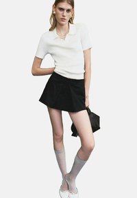 Mujer joven con una blusa blanca de manga corta, falda corta negra, calcetas blancas hasta la rodilla, bailarinas blancas y sosteniendo un bolso negro.