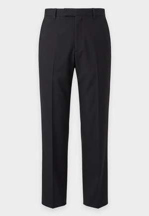 Homme en polo blanc, pantalon noir et chaussures noires tenant un grand sac cabas en cuir noir sur un fond uni clair.