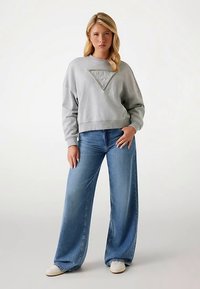 Sweat-shirt gris avec un logo triangulaire sur la poitrine, associé à un jean denim bleu clair à jambes larges et des baskets blanches.