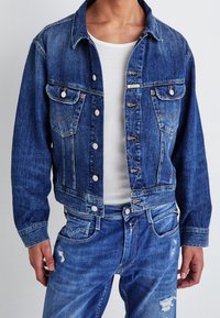 Man wearing blue denim jacket over white ribbed tank top and distressed blue jeans, standing against plain background.