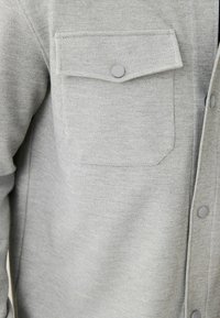 Camicia grigia con bottoni realizzata in tessuto testurizzato, dotata di una tasca sul petto con pattina arrotondata, dettagli di cucitura sottili e bottoni grigi.