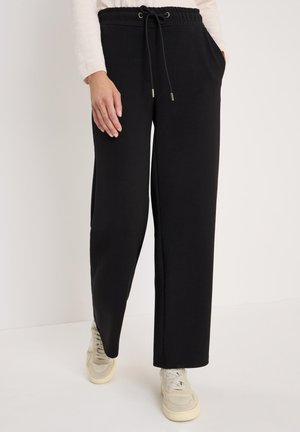 Pantalons de survêtement noirs avec ceinture élastique, cordon de serrage ajustable, poches latérales et coupe décontractée. Associés à des baskets grises.