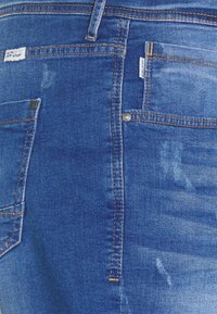 Vaqueros de mezclilla en azul medio, con costuras en contraste, bolsillos delanteros y traseros, y una textura desgastada con parches ligeramente descoloridos.