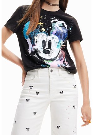 Vrouw draagt een zwart T-shirt met een kleurrijk Mickey Mouse gezicht en witte broek met kleine bedrukt Mickey Mouse gezichtjes.