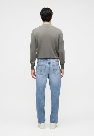 Άνδρας με την πλάτη στραμμένη, φορώντας γκρι μπλούζα μακρυμάνικη, ανοιχτόχρωμα τζιν με πίσω τσέπες και λευκά αθλητικά παπούτσια, σε απλό φόντο.