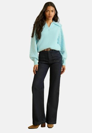 Hellblauer Strickpullover mit V-Ausschnitt und Puffärmeln, kombiniert mit dunklen ausgestellten Jeans. Ohrringe und ein brauner Gürtel vervollständigen den Look.