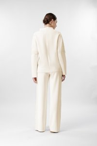 Mujer con el cabello recogido en un moño, vestida con una blusa de manga larga color crema y pantalones de pierna ancha, de pie de espaldas contra un fondo liso.