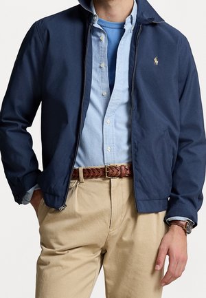 Mann trägt eine marineblaue Jacke über einem hellblauen Hemd mit Knöpfen und einem blauen T-Shirt, beige Hose, braunen geflochtenen Gürtel und Armbanduhr am linken Handgelenk.