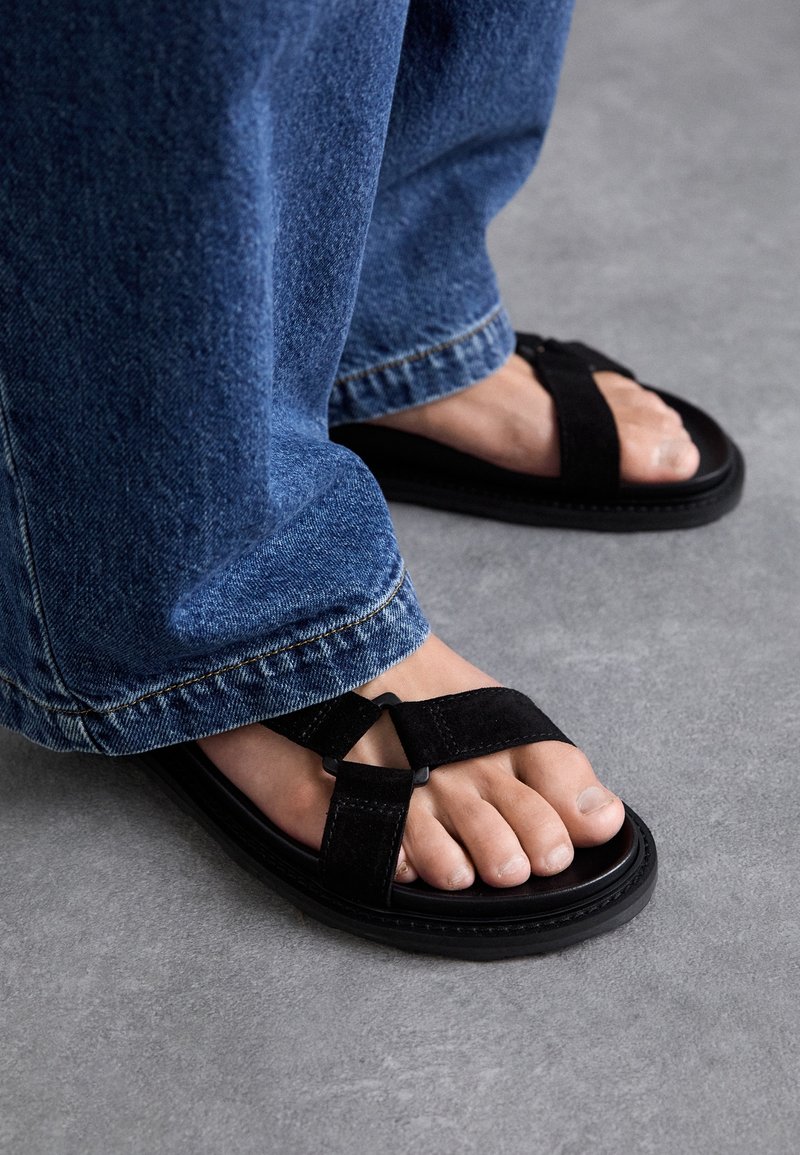 Pieds portant des sandales noires avec de larges brides en tissu et un jean en denim bleu debout sur un sol gris.