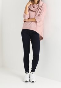 Kobieta ubrana w różowy sweter z wysokim kołnierzem, czarne obcisłe spodnie i białe trampki, stoi z rękami skrzyżowanymi wzdłuż białej ściany.