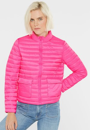 Frau mit kurzem blonden Haar, die eine knallpinke gesteppte Jacke und eine blaue Jeans trägt, steht mit einer Hand in die Hüfte gestützt.