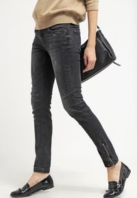 Jeans skinny negros con un acabado desgastado, con detalles con cremalleras en los tobillos. Llevados con un suéter beige y zapatos planos negros.