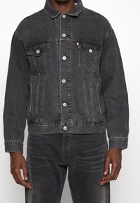 Svart denimjacka med en klassisk design, två bröstfickor, en spetsig krage och tryckknappar. Har en subtil regnbågsetikett nära fickan.