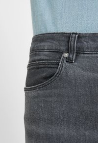 Jeans de mezclilla gris oscuro con un corte ajustado, que presentan un bolsillo frontal, detalles cosidos y una pretina estándar. La textura parece suave.